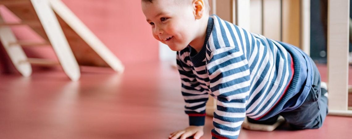 A cheerful toddler in striped long sleeves crawls on a red floor indoors, radiating joy.